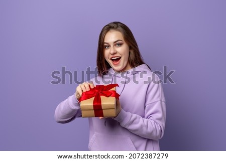 Happy amazed brunet woman in purple hoodie opening a present, standing over purple background. New Year Women's Day birthday holiday concept