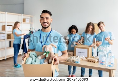 Arab male volunteer holding box with clothes and toys for donation, smiling to camera while working at charity center with group of colleagues, packing products in boxes on background Royalty-Free Stock Photo #2072405393