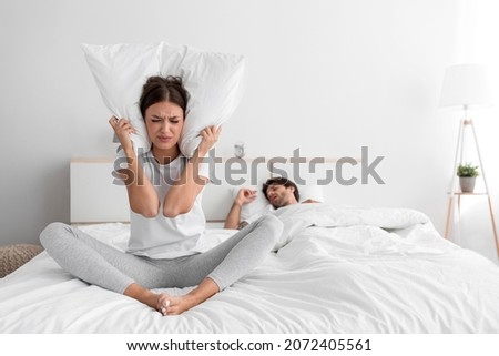 Stressed sad millennial caucasian lady sits on bed covers ears with pillow and suffers from snoring of sleeping man at night, empty space. Insomnia, sleep problems, apnea and relationship difficulties Royalty-Free Stock Photo #2072405561