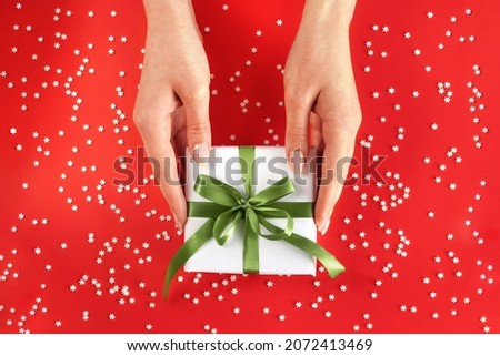 Nice female hands holding gift box wrapped in white paper with green ribbon on festive red background with many snowflakes. Xmas and New Year postcard design. Giving love and warmth, Christmas concept