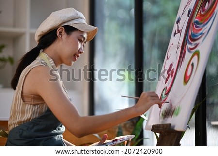 Happy young asian woman painting picture on canvas with oil paints in her studio.