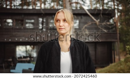 Portrait of a Young Beautiful Blond Woman in a Romantic Nature Atmosphere with a Wooden Forest Cottage in the Background. She is Confident and Expresses a Light Smile.