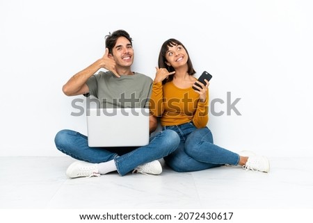 Young couple with a laptop and mobile sitting on the floor making phone gesture. Call me back sign