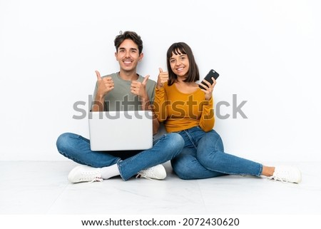 Young couple with a laptop and mobile sitting on the floor giving a thumbs up gesture with both hands and smiling