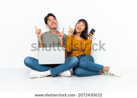 Young couple with a laptop and mobile sitting on the floor pointing with the index finger a great idea