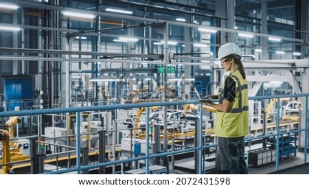 Female Car Factory Engineer in High Visibility Vest Using Laptop Computer. Automotive Industrial Manufacturing Facility Working on Vehicle Production with Robotic Arms. Automated Assembly Plant. Royalty-Free Stock Photo #2072431598