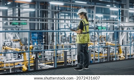 Female Car Factory Engineer in High Visibility Vest Using Laptop Computer. Automotive Industrial Manufacturing Facility Working on Vehicle Production with Robotic Technology. Automated Assembly Plant. Royalty-Free Stock Photo #2072431607