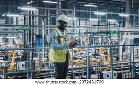 Happy African American Car Factory Engineer in High Visibility Vest Using Tablet Computer. Automotive Industrial Facility Working on Vehicle Production on Automated Technology Assembly Plant. Royalty-Free Stock Photo #2072431700