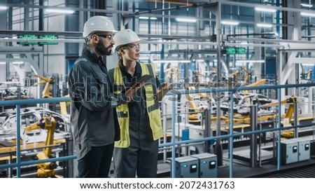 Male Specialist and Female Car Factory Engineer in High Visibility Vests Using Tablet Computer. Automotive Industrial Manufacturing Facility Working on Vehicle Production. Diversity on Assembly Plant. Royalty-Free Stock Photo #2072431763