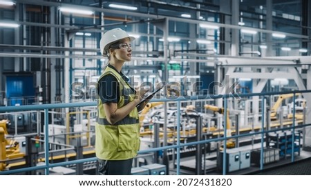 Female Car Factory Engineer in High Visibility Vest Using Laptop Computer. Automotive Industrial Manufacturing Facility Working on Vehicle Production with Robotic Arms. Automated Assembly Plant. Royalty-Free Stock Photo #2072431820