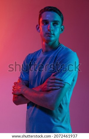 Motivated, composed. Portrat of Caucasian young sportive man in sportswear standing isolated on pink background with blue neon filter, light. Concept of action, motion, speed, healthy lifestyle.