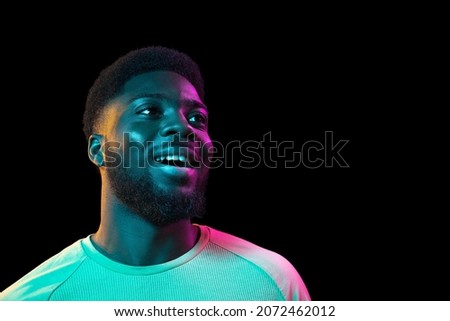 Smiling looking at side. African young man's portrait on dark studio background in neon light. Beautiful male model in casual style. Concept of human emotions, facial expression, youth, sales, ad.