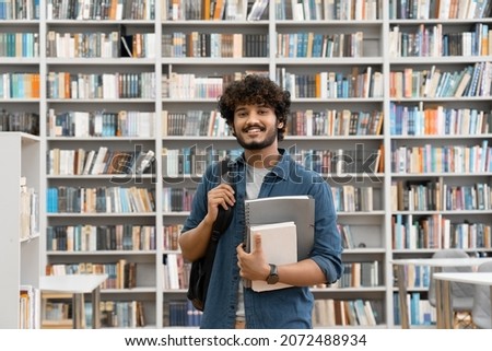 Young smiling Indian male student standing in university library with backpack and studying accessories. Guy bookworm looking at camera with confident smile over book store interior portrait