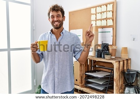 Young handsome man drinking a cup coffee at the office smiling happy pointing with hand and finger to the side 