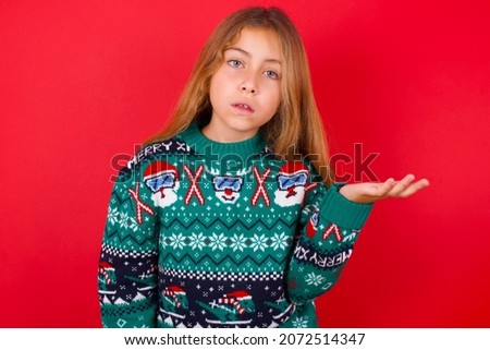 Studio shot of frustrated brunette kid girl in knitted sweater christmas over red background gesturing with raised palm, frowning, being displeased and confused with dumb question.