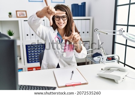 Young doctor woman wearing doctor uniform and stethoscope at the clinic smiling making frame with hands and fingers with happy face. creativity and photography concept. 
