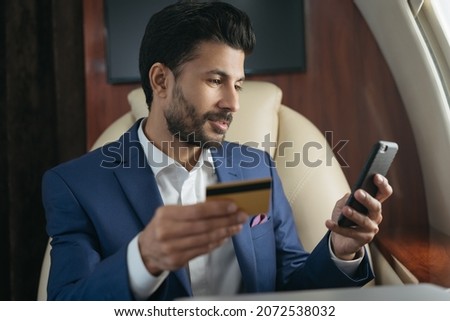 Handsome Arabian businessman holding credit card using  mobile phone, shopping online sitting in airplane. Confident middle eastern entrepreneur receive payment, flying luxury private jet