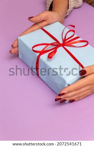 Female hands hold wrapped gift with red ribbon. Purple background with place for text. Advertising of gifts. Selective focus.
