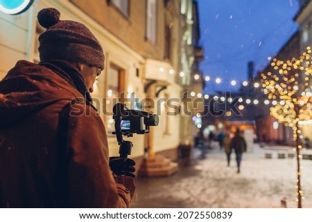 Videographer filming Christmas lights on city street at night. Man using steadicam and camera for footage under falling snow. New year video shoot. Digital devices
