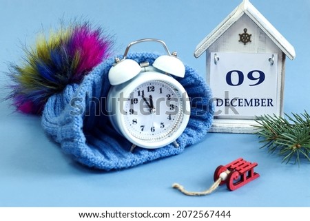 Calendar for December 9: decorative house with the name of the month in English, numbers 09, a winter blue hat, a clock in it, fir branches and toys, blue background