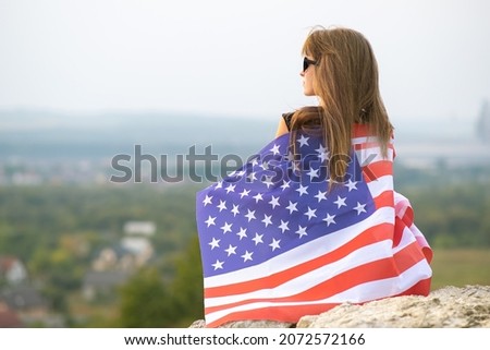 Young pretty american woman with long hair holding waving on wind USA  flag on her sholders resting outdoors enjoying warm summer day.