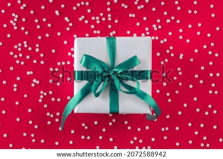 Gift box wrapped in white paper with a mint blue bow on festive crimson background with many snowflakes. Copyspace for your text. Flat lay style. Christmas, New Year or birthday celebration concept
