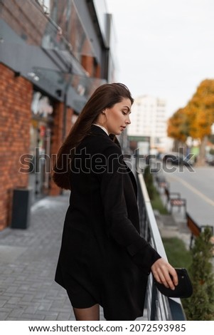 Fashionable beautiful young girl with a purse in black business clothes with a coat and skirt walking in the city 