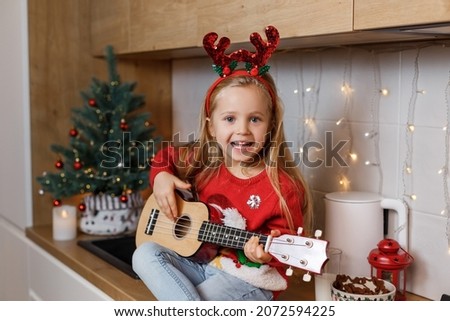 Happy little girl in red horns playing ukulele on the background of christmas tree. Talent kid having fun on winter holidays at cozy decorated home kitchen. Royalty-Free Stock Photo #2072594225