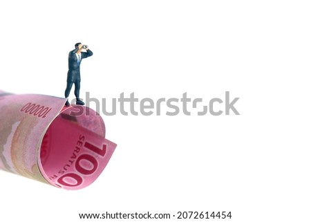 Businessman using binoculars standing above rolled money paper. Miniature tiny people toys photography. isolated on white background