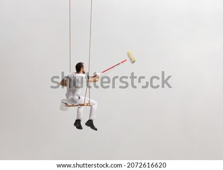 Painter sitting on a swing and painting a wall isolated on white background Royalty-Free Stock Photo #2072616620