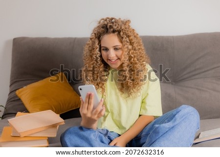 blogger girl with headphones and a smartphone in her hands is sitting cross-legged on a gray sofa. young woman with curly hair communicates online. High quality photo