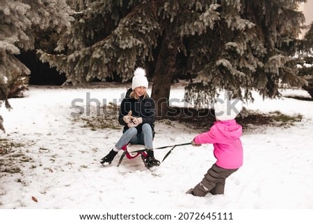 A little daughter rolls her mother on a sled in the snow.