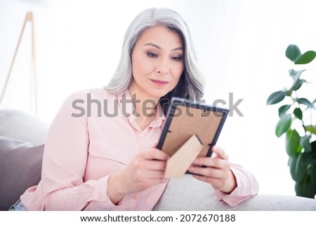 Photo of attractive aged korean woman happy smile look photo frame nostalgia remember sit couch home