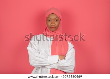 Arab woman with serious and calm expression with dark skin, wrapped in pink veil, wears white shirt on pink studio wall.