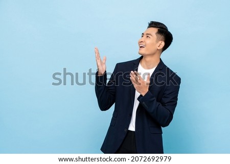 Young smiling Asian man opening hands and looking up sideways in isolated light blue color background Royalty-Free Stock Photo #2072692979