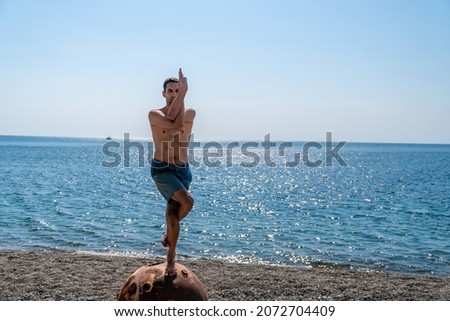 Man doing yoga exercise outdoors standing on old rusty floating marine mine on the beach with rocky shore and sea background. Healthy lifestyle, pollution, nature protection, war and peace concept.