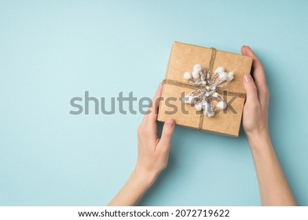 First person pov top view photo of woman's hands holding craft paper giftbox decorated with snow branch and twine on isolated pastel blue background with copyspace
