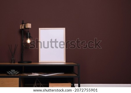 Empty frame and stylish lamp on wooden table near brown wall. Mockup for design