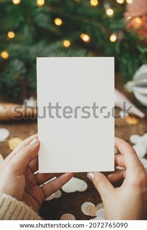 Hands holding empty greeting card and christmas ornaments, confetti, pine branches and lights on rustic wooden background. Christmas card mock up. Space for text. Seasons greetings. Template