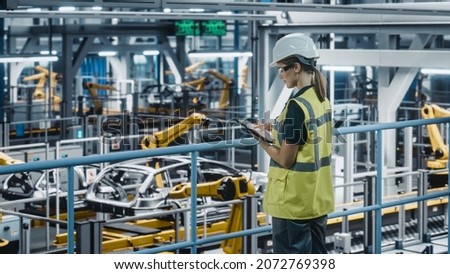 Female Car Factory Engineer in High Visibility Vest Using Tablet Computer. Automotive Industrial Manufacturing Facility Working on Vehicle Production with Robotic Technology. Automated Assembly Plant. Royalty-Free Stock Photo #2072769398