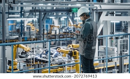 Multiethnic Car Factory Engineer in Work Uniform Using Tablet Computer. Automotive Industrial Manufacturing Facility Working on Vehicle Production with Robotic Arms. Automated Assembly Plant. Royalty-Free Stock Photo #2072769413