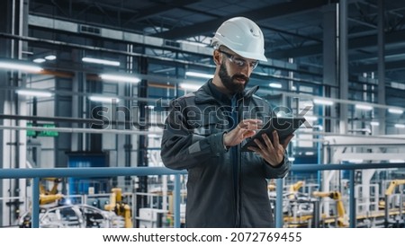 Portrait of Automotive Industry Engineer in Safety Glasses and Uniform Using Laptop at Car Factory Facility. Professional Assembly Plant Specialist Working on Manufacturing Modern Electric Vehicles.