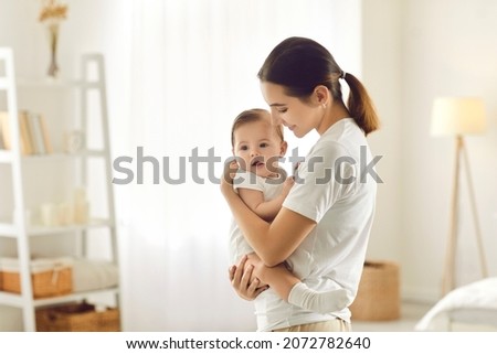 Mother holding baby. Happy beautiful young mom standing in bedroom or nursery room interior in tender white and pastel colors, holding cute sweet little child and smiling. Family, love, care concept
