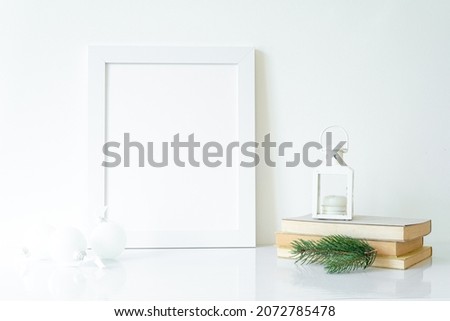Vertical white blank frame mockup with Christmas lantern, books, fir tree branch and white Christmas decorations. Christmas, winter, festive concept. Copy space
