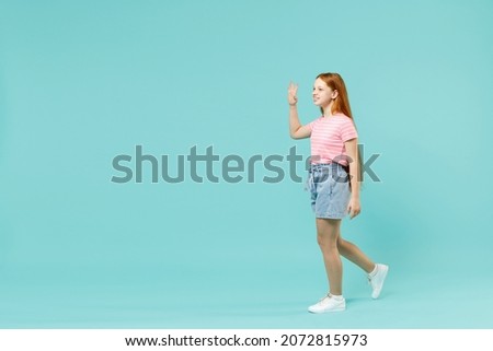 Full length little smiling happy redhead kid girl 12-13 years old in pink striped t-shirt hold walking going waving hand isolated on pastel blue background studio. Children lifestyle childhood concept