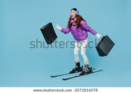 Full body happy skier woman in warm purple padded windbreaker jacket ski goggles mask spend weekend in mountains holding package bags with purchases shopping isolated on plain blue background studio