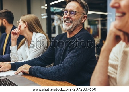 Mature businessman attending a meeting with his colleagues. Happy mature businessman laughing cheerfully while sitting in a boardroom with his team. Group of colleagues working together in an office. Royalty-Free Stock Photo #2072837660