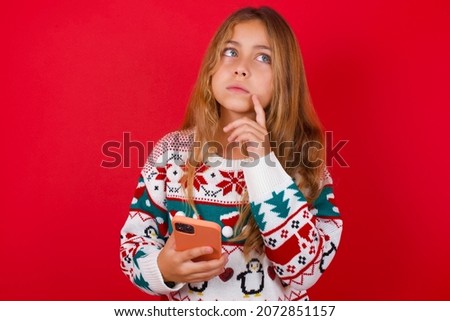 brunette kid girl in knitted sweater christmas over red background thinks deeply about something, uses modern mobile phone, tries to made up good message, keeps index finger near lips.