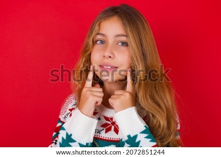 Happy brunette kid girl in knitted sweater christmas over red background with toothy smile, keeps index fingers near mouth, fingers pointing and forcing cheerful smile