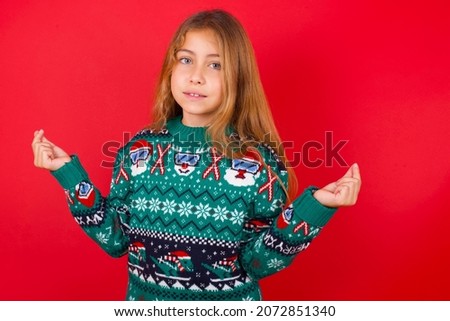 brunette kid girl in knitted sweater christmas over red background , making money gesture.